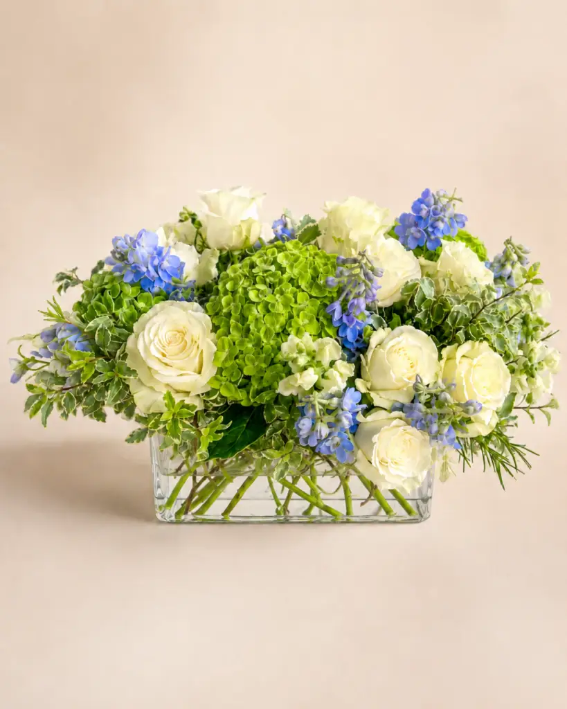 Ocean Meadow floral centerpiece with white roses, green hydrangea, soft blue blooms, white seasonal flowers, variegated greenery, and textural greens in a low glass vase by a Santa Monica florist.